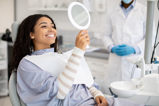 Person seated in a dental chair smiles into a handheld mirror, wearing a striped shirt and dental bib. A masked dental professional stands nearby in a modern clinic, suggesting a successful check-up or procedure.