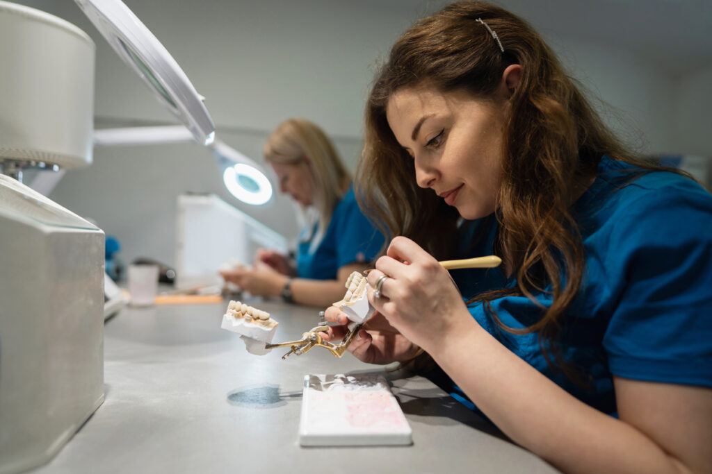 Two individuals work in a dental lab, one painting a dental mold with a small brush under bright lighting. Another person in the background uses a magnifying lamp, highlighting the precision and craftsmanship of dental prosthetic creation.