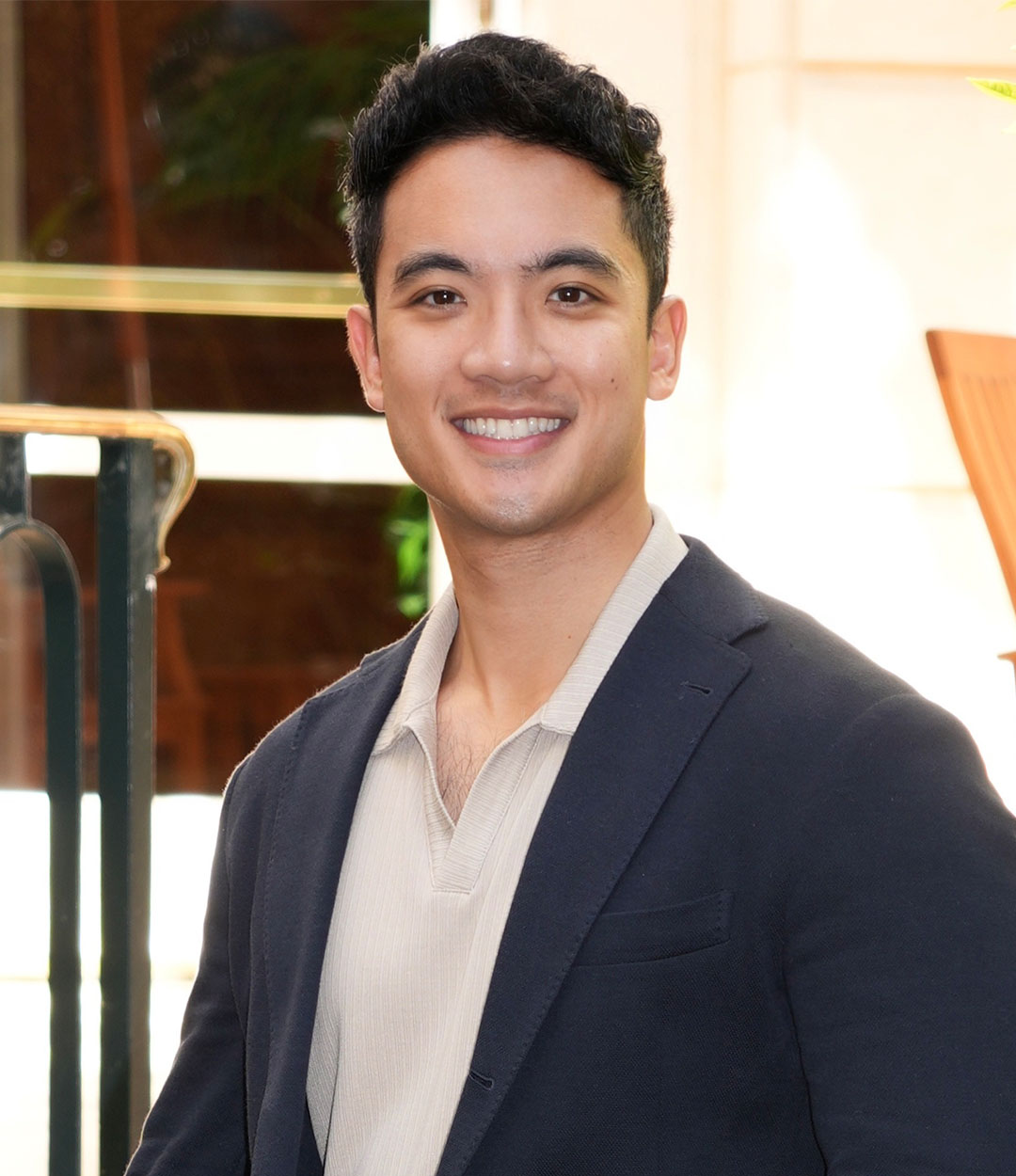 Headshot of Dr. Geoff Mateo, DMD with short dark hair wears a dark blazer over a light shirt, standing indoors with a blurred background. Soft lighting and visible plant elements suggest a professional yet relaxed setting.